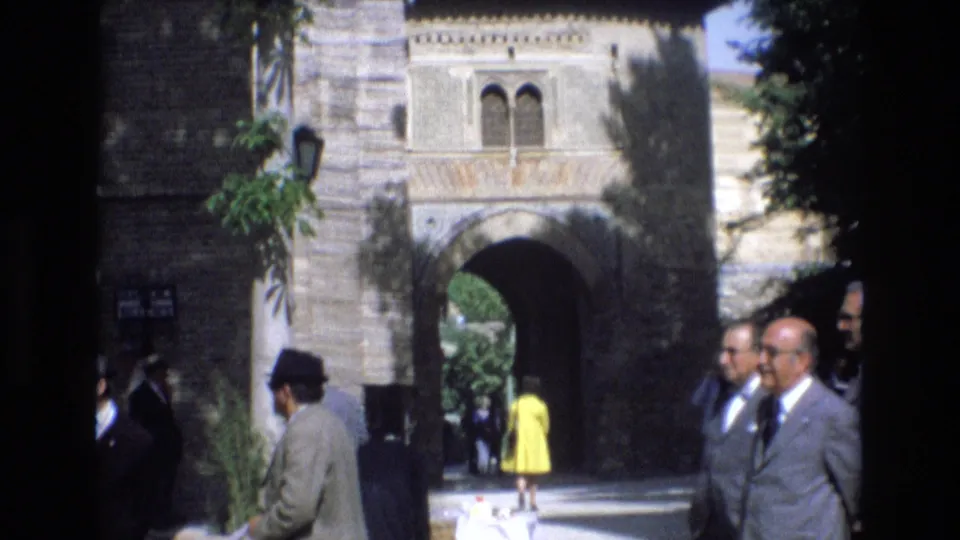 Visitors in suits and bright clothing move near an ornate stone archway in Spain, filmed from a handheld 8mm travel perspective.