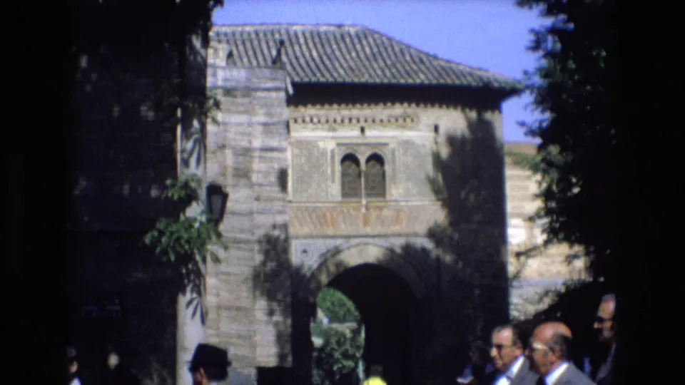 The final frame shows a historic stone gateway with decorative masonry and roof tiles as visitors stand and walk near the shaded entrance.