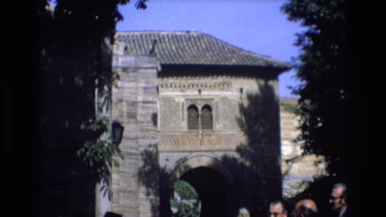 1972 Spain home-movie frame showing a historic-looking stone archway with arched windows, tiled roof, tree shadows, and adult men gathered along the lower edge.