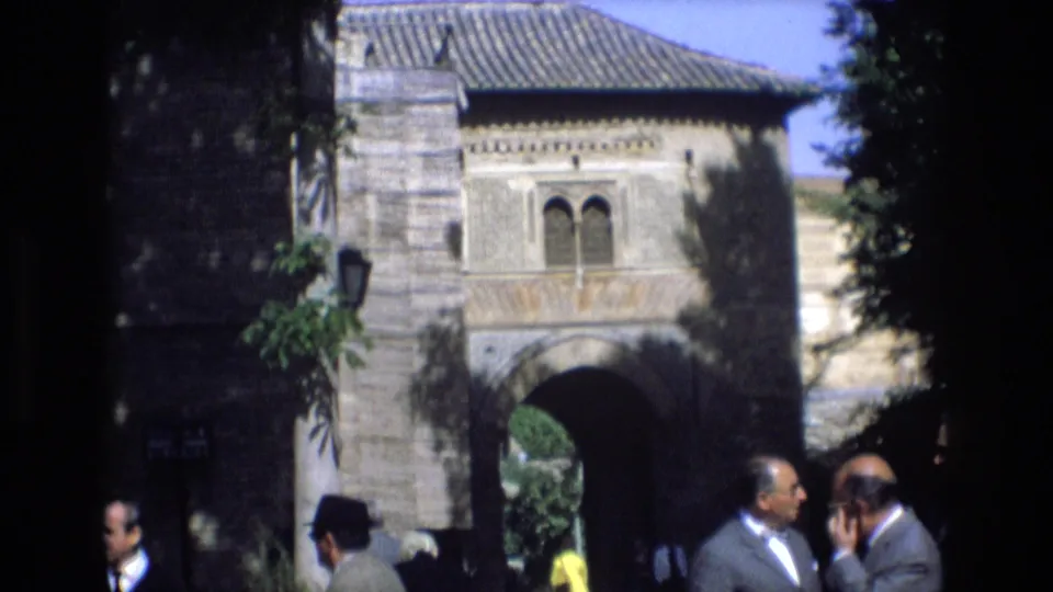 1972 Spain home-movie still showing a closer visitor-shot view of the stone archway, decorative upper window, hanging foliage, and men conversing in the foreground.