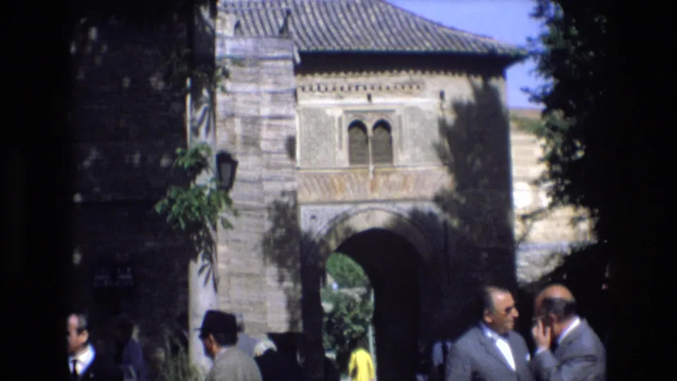 1972 Spain frame showing the same historic-looking archway with a dark shaded foreground, adult men along the bottom edge, and bright greenery visible through the opening.