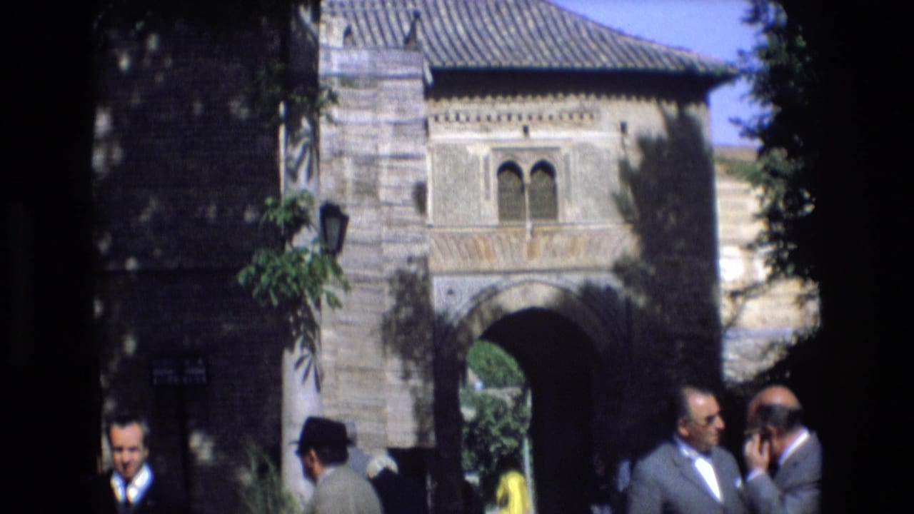 1972 Spain 8mm frame showing a historic stone gateway with arched windows above and adult visitors in suits and hats near the entrance.