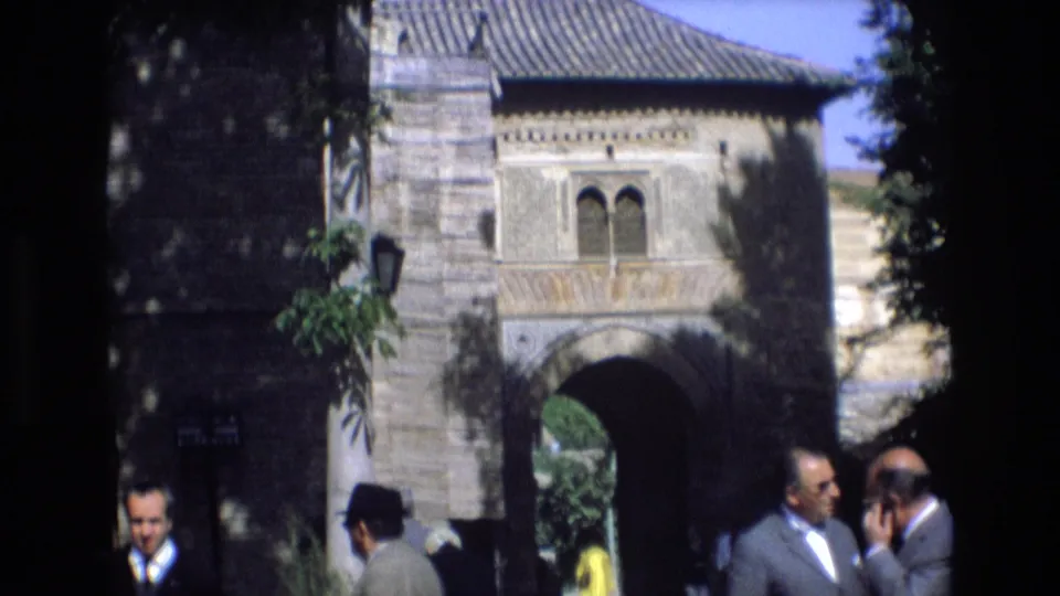 1972 Spain 8mm frame showing a historic stone gateway with arched windows above and adult visitors in suits and hats near the entrance.
