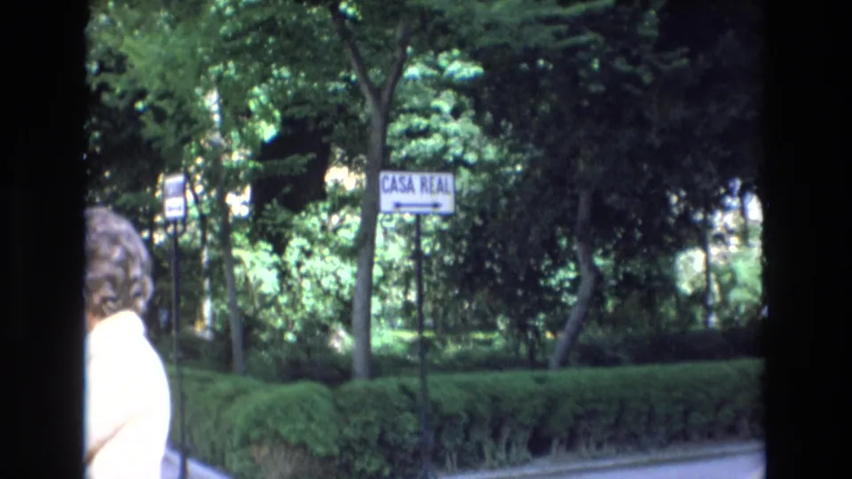 1972 Spain 8mm frame showing a green park or estate edge with hedges, trees, and a roadside sign reading "CASA REAL," with an adult partly visible in the foreground.