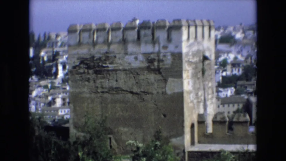 1972 Spain 8mm frame showing a weathered stone turret and adjoining battlements with white buildings on the hillside behind.