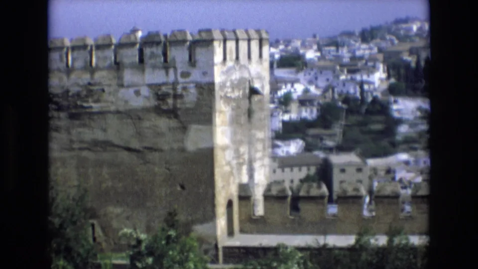 1972 Spain 8mm frame showing a fortress wall and crenellated turret in the foreground with rooftops and a hillside town beyond.