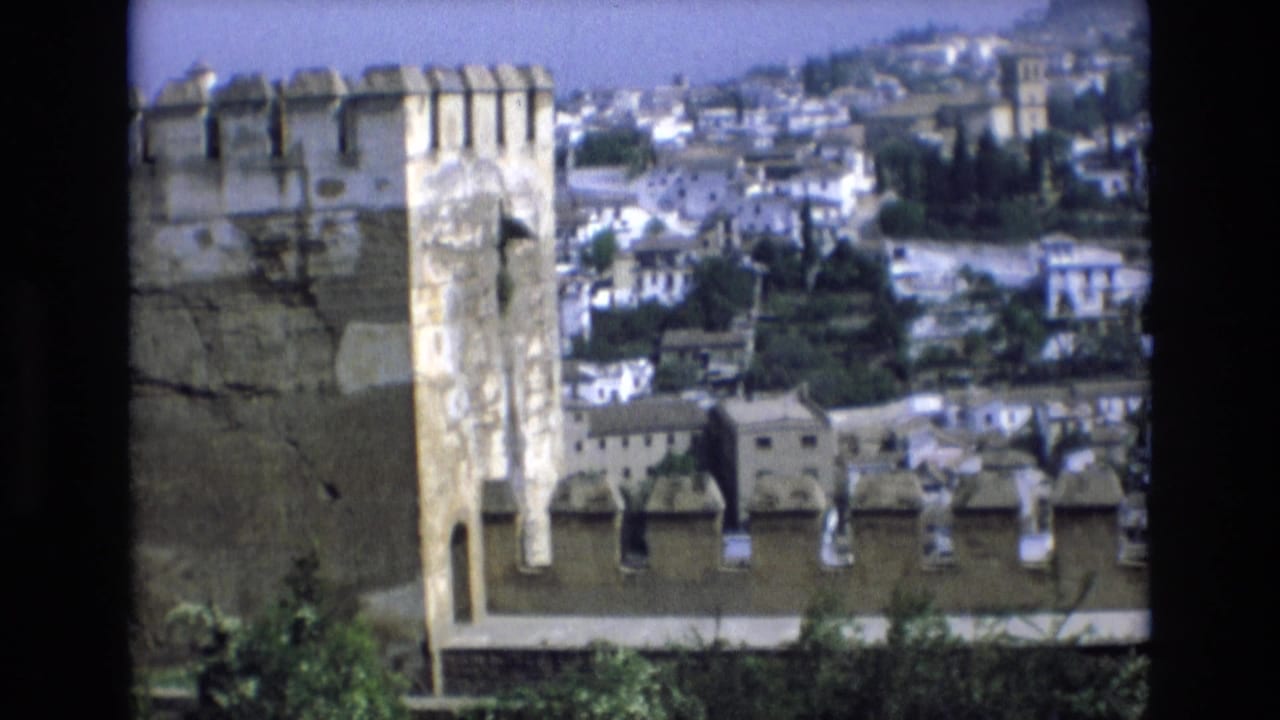 1972 Spain 8mm frame showing a stone fortress tower and crenellated wall in front of white hillside buildings and trees.