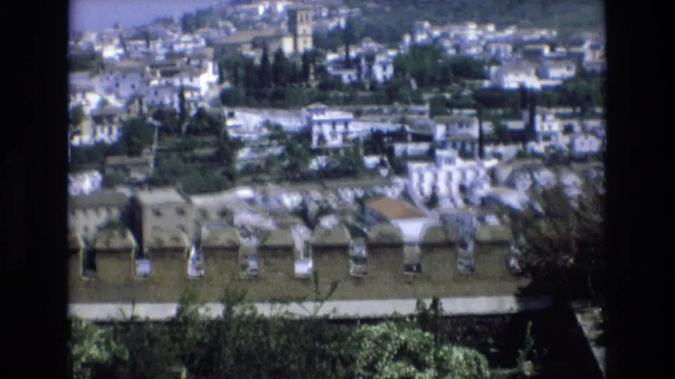 1972 Spain 8mm frame panning across crenellations, rooftops, white buildings, vegetation, and a distant tower in a historic townscape.