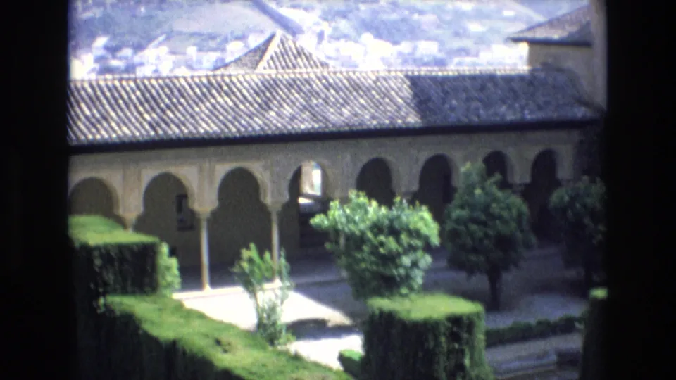 1972 Spain 8mm frame showing a quiet courtyard with arched colonnades, tiled roofing, clipped hedges, shrubs, and paved walkways.