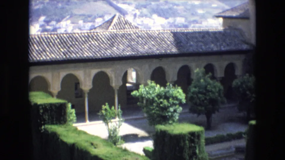 1972 Spain 8mm frame showing a Moorish-style arched arcade above a formal courtyard garden with hedges, paths, and small trees, seen from a dark window or balcony edge.