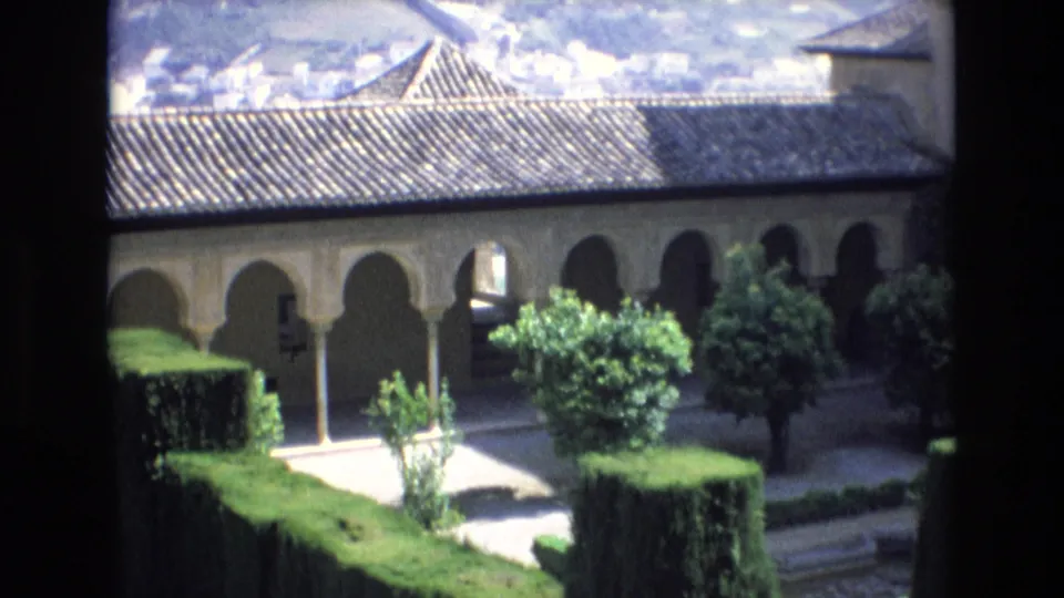 Soft 8mm frame from Spain in 1972 showing a sunlit courtyard garden, clipped hedges, small ornamental trees, and a shaded arched facade with a tiled roof.