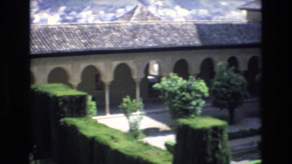 1972 Spain 8mm frame showing the courtyard from an elevated position, with dark borders framing a tiled-roof arcade, manicured hedges, trees, and pale walkways.