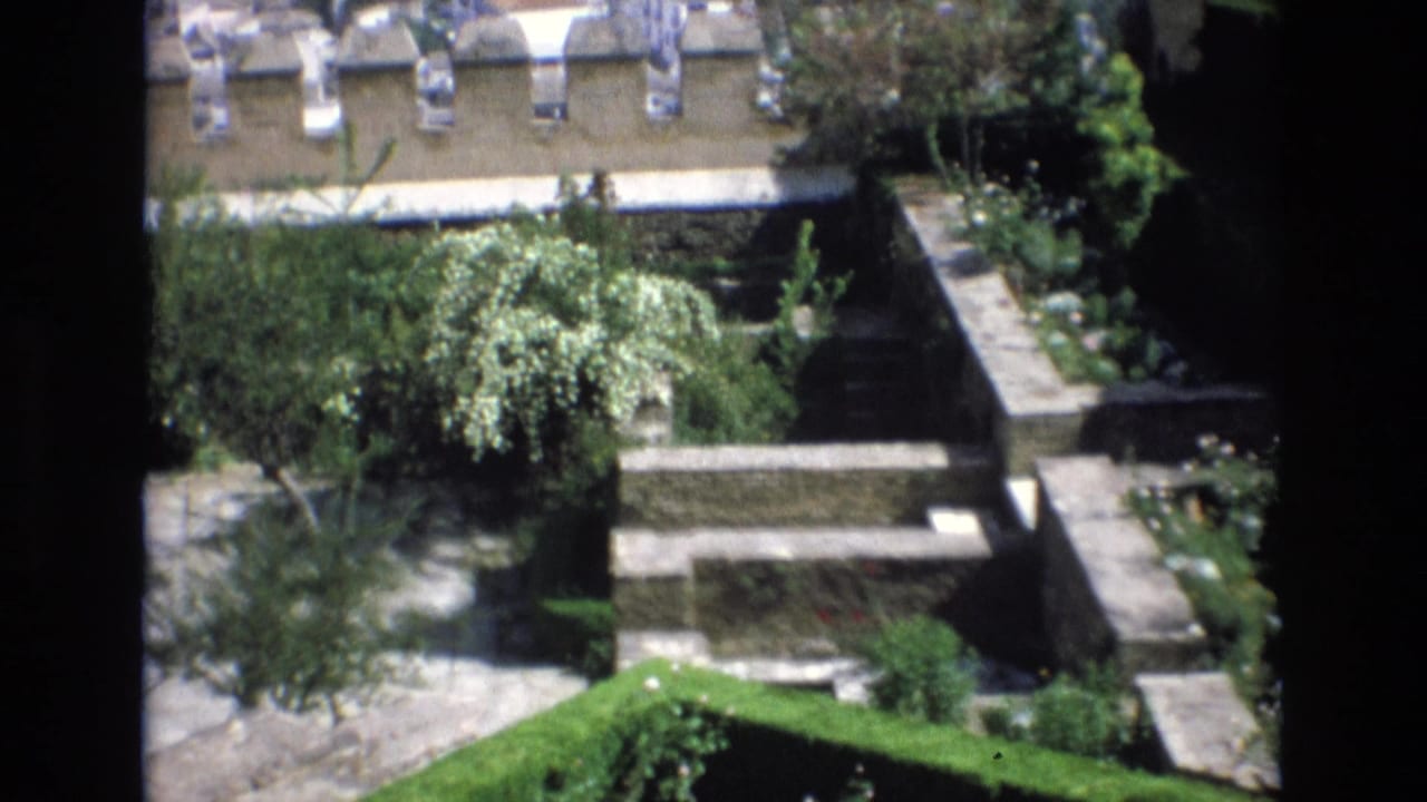 1972 Spain home-movie frame showing a sunlit terraced courtyard with stone retaining walls, garden trees, and tiled rooftops in the background.