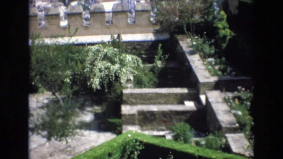 1972 Spain home-movie frame showing a sunlit terraced courtyard with stone retaining walls, garden trees, and tiled rooftops in the background.