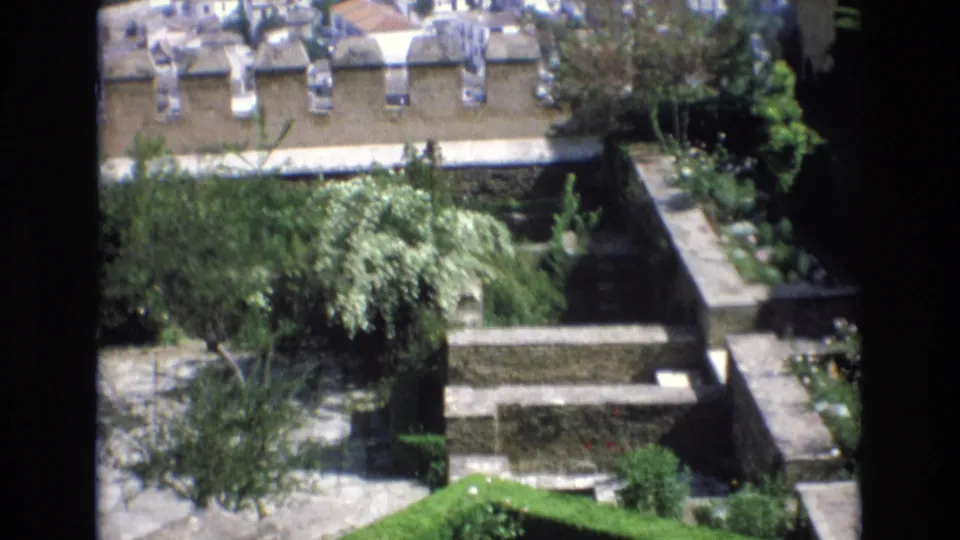 1972 Spain home-movie view of a terraced stone courtyard with garden trees, low walls, paths, and a bright green lawn edge in sunlight.