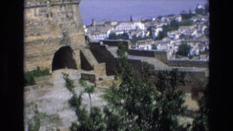 1972 Spain frame showing a stone terrace with an arched masonry opening and a distant town of white buildings under bright daylight.