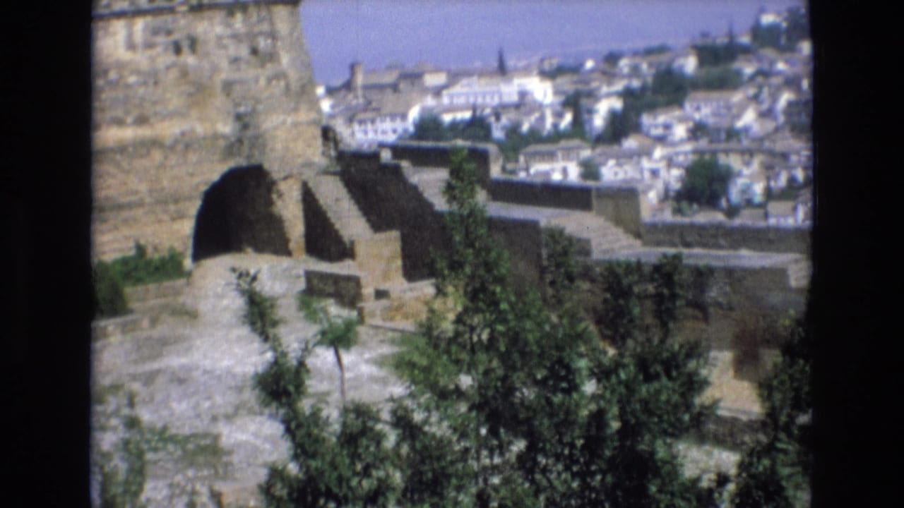 1972 Spain 8mm frame showing a weathered stone fortification with an arched opening, foreground trees, and a distant white old-city view.