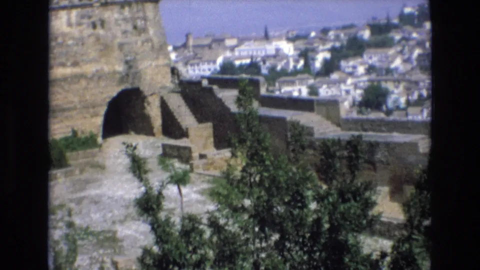 1972 Spain 8mm frame showing a weathered stone fortification with an arched opening, foreground trees, and a distant white old-city view.