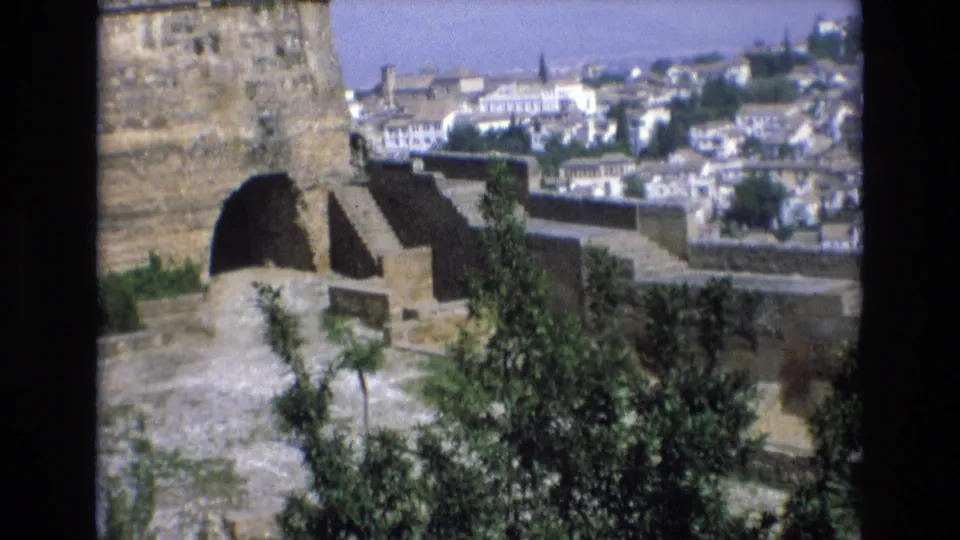 1972 Spain 8mm frame of a stone fortress terrace with an arched recess, green foliage, and white buildings spread across the background.