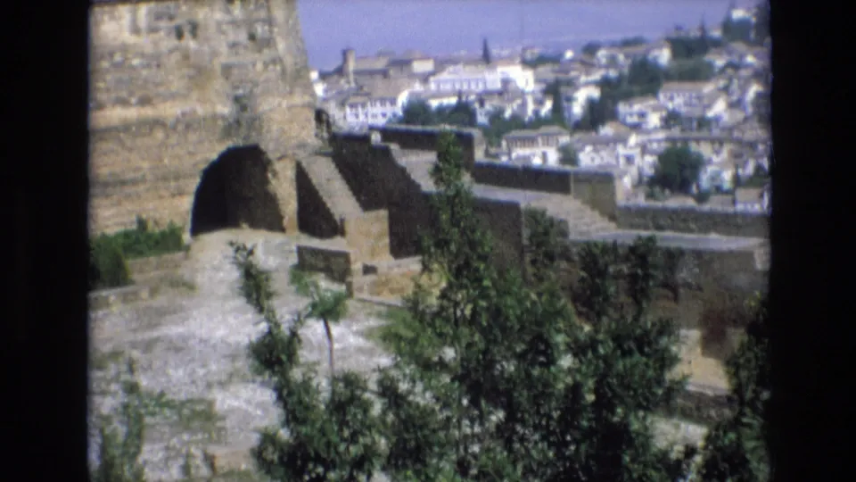 1972 Spain 8mm frame showing fortress masonry and a paved overlook with trees in front of a sunlit old city.