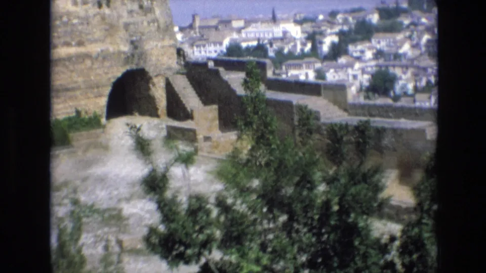 1972 Spain 8mm frame with leafy foreground branches, a stone arched structure, and white old-city buildings visible beyond the wall.