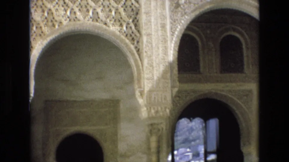 1972 Spain 8mm frame with carved archways, patterned stucco panels, upper arched windows, and small distant visitor silhouettes near a bright opening.