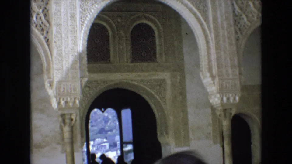 1972 Spain 8mm frame of an ornate palace arch and carved wall details, with a bright exterior view and small visitor silhouettes visible through the opening.
