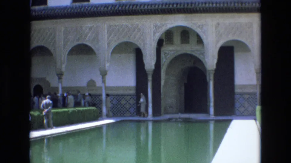 1972 Spain 8mm frame widening to a palace courtyard with a reflective green pool, arched colonnade, tiled wall band, and visitors standing along the walkway.