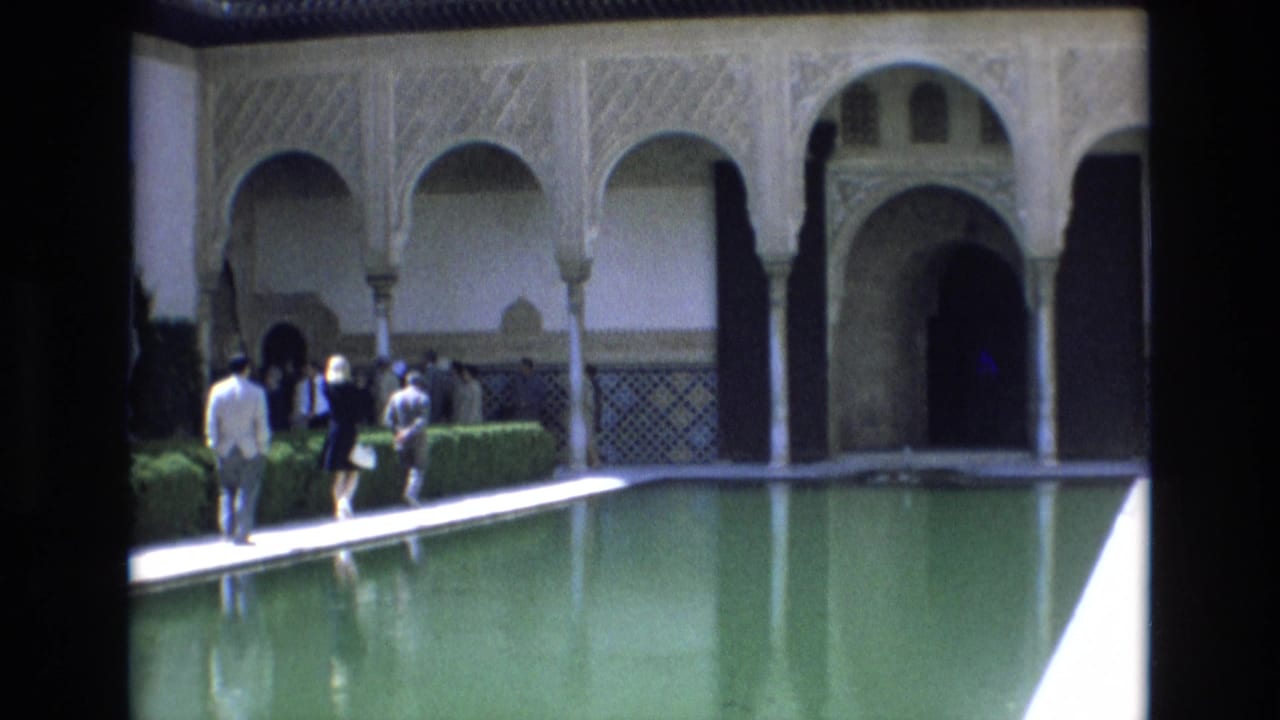 Several visitors stand and walk along a pool edge below arched architecture.