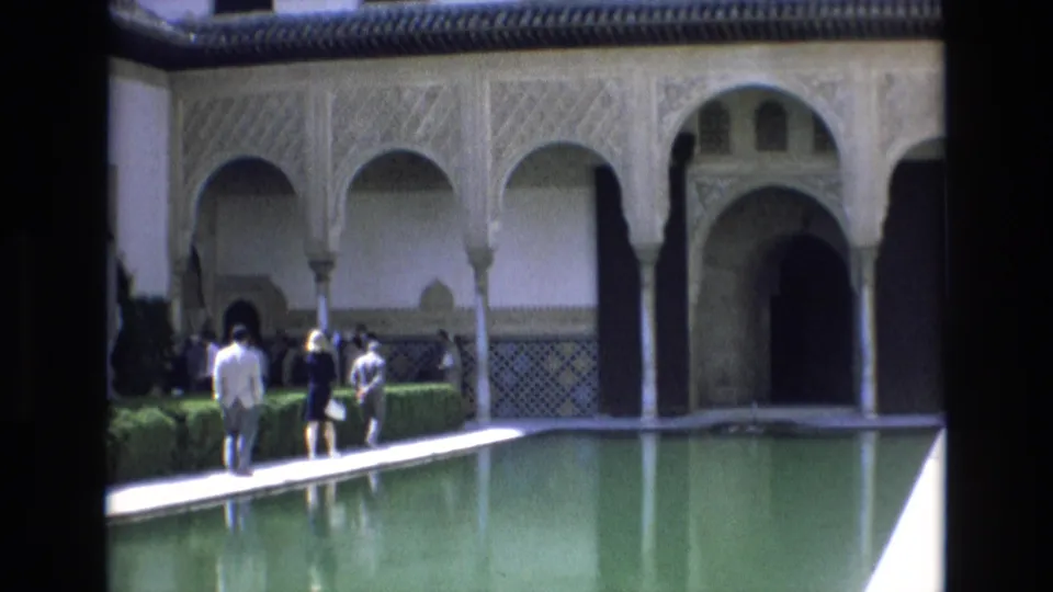 A visitor in a light jacket stands near the reflecting pool as other adults walk through the historic courtyard in Spain.