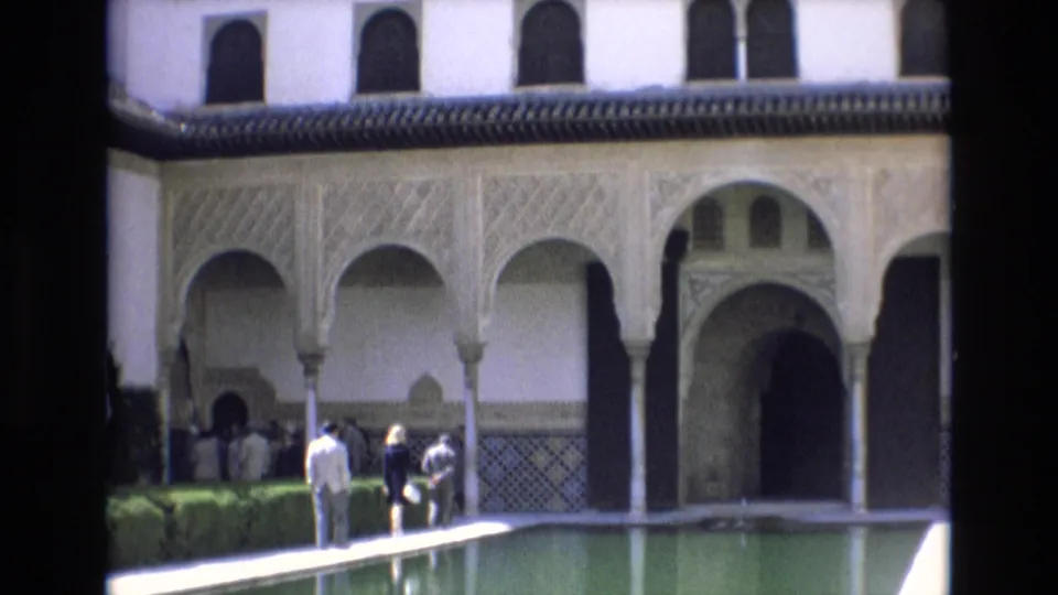 A fixed 8mm view shows visitors beside the reflecting pool, framed by carved arches, patterned tilework, hedges, and a dark courtyard doorway.