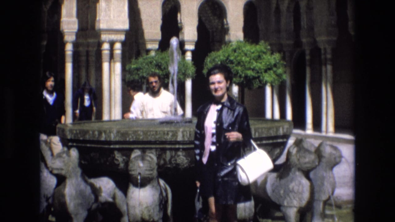 In Spain in 1972, a woman in a dark jacket and pink top poses beside a stone fountain with sculptural animal forms, arched colonnades, clipped trees, and tourists in the background.