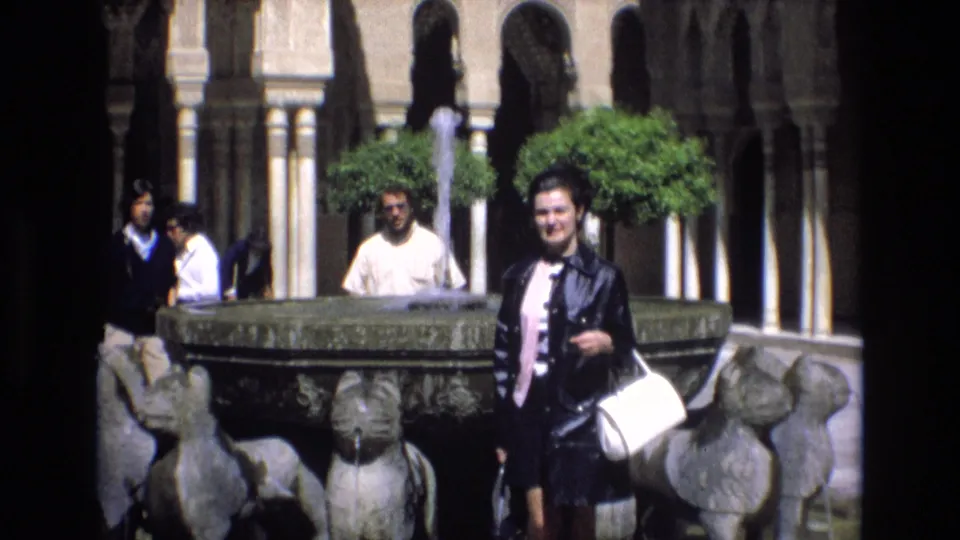 The woman remains near the ornate fountain in a Spanish courtyard, with running water, clipped trees, arched columns, and several visitors visible behind her.
