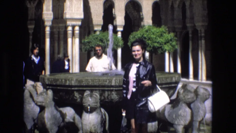 A handheld 1972 Spain travel frame shows the woman by the sculptural fountain, with tourists, arched courtyard walls, trees, and water spray visible around the basin.