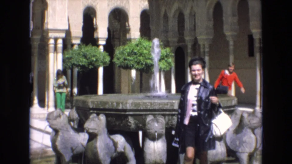 At the end of the clip, the woman stands at the right side of the fountain, with sculptural supports, arched architecture, clipped trees, water, and passing visitors visible in the courtyard.