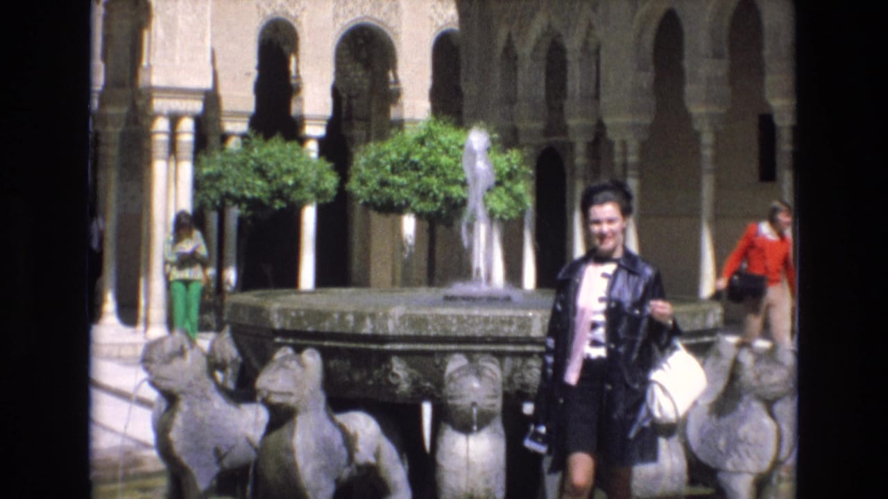 In Spain in 1972, a woman in a black jacket and pink top smiles toward the camera beside a stone fountain with sculpted animal heads in a courtyard with arches and trimmed trees.