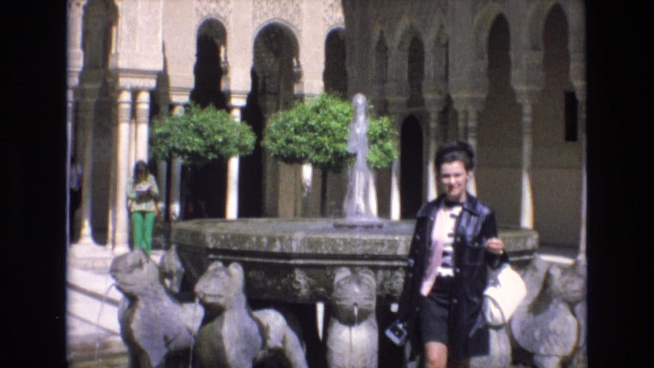 Woman stands near a fountain with arched colonnades behind her.