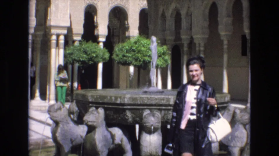 A woman in 1972 Spain poses near the fountain edge, with carved animal-head forms in the foreground and a visitor in green pants visible beneath the courtyard arches.