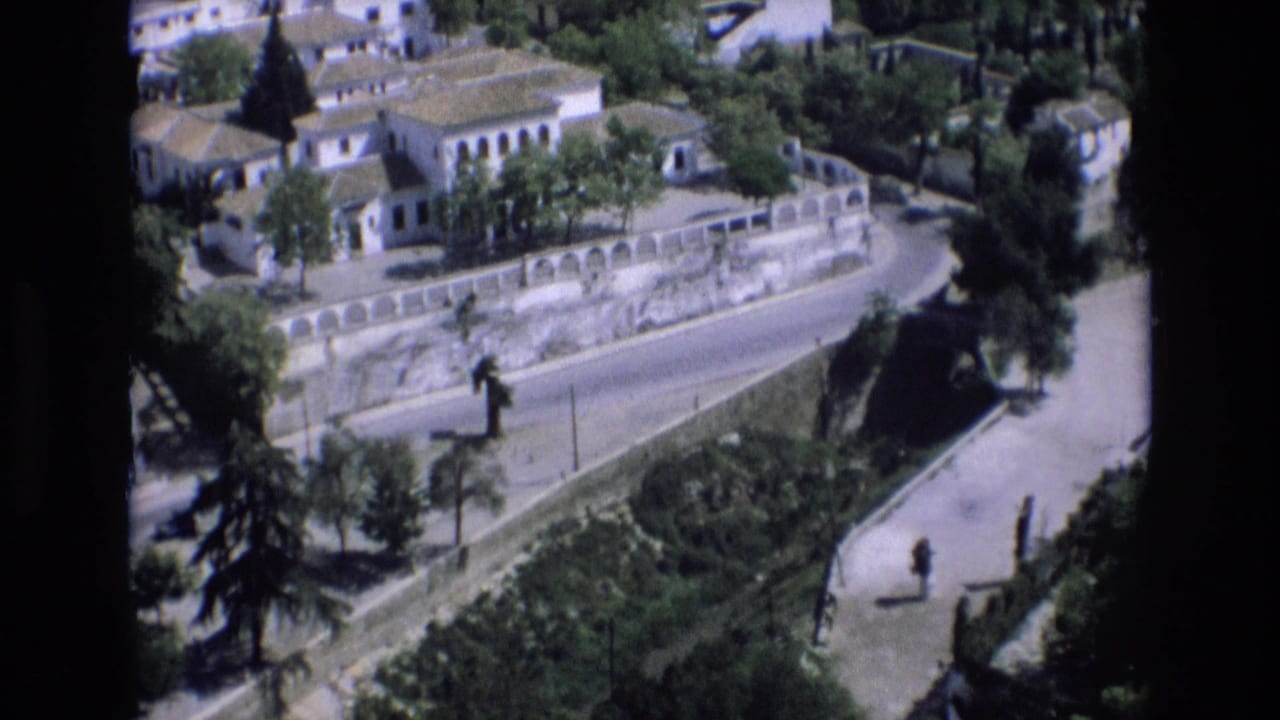 1972 Spain home-movie frame showing an elevated view over white buildings, terracotta roofs, trees, a curved stone wall, and a quiet road.