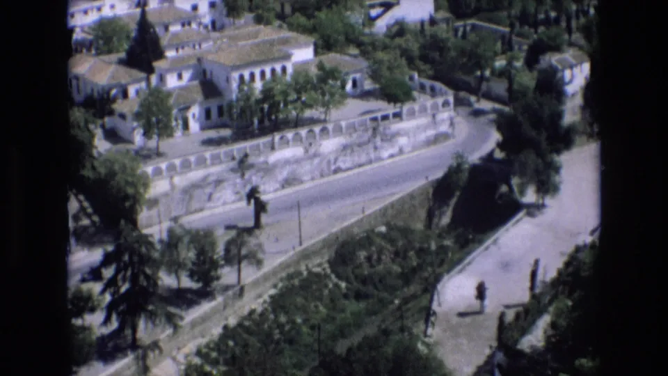1972 Spain home-movie frame showing an elevated view over white buildings, terracotta roofs, trees, a curved stone wall, and a quiet road.