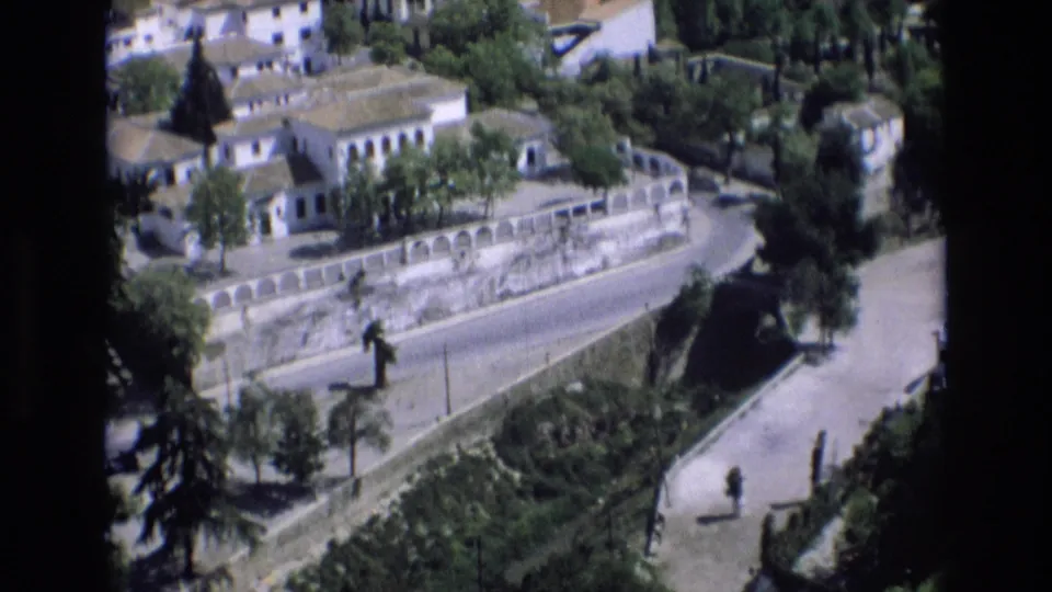 1972 Spain frame with whitewashed buildings among trees above a long stone retaining wall, with a winding road and small pedestrian below.