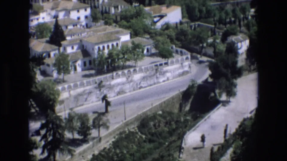 Elevated 1972 Spain home-movie view of town buildings, a long stone wall, road curves, trees, and small distant pedestrians.