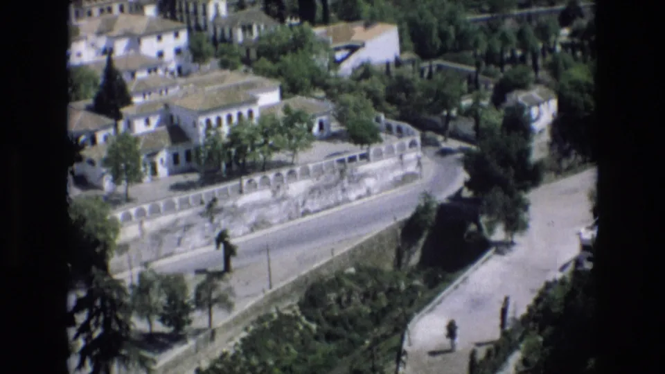 1972 Spain frame showing a high-angle view of white buildings, tree cover, a curved stone wall, a road, and a pedestrian pathway.