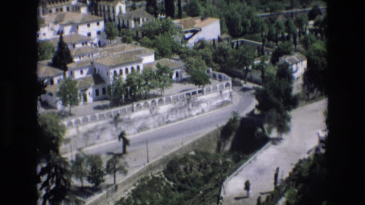 Elevated 1972 Spain home-movie view of pale town buildings, terracotta roofs, trees, and a curved road bordered by a stone retaining wall.