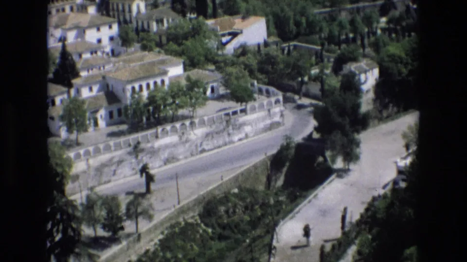 Elevated 1972 Spain view across white and pale buildings, trees, a low stone wall, and a quiet paved road or plaza edge.
