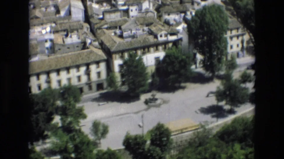 Elevated 1972 Spain home-movie frame showing tiled rooftops, pale facades, a tree-shaded plaza, and a small central fountain.