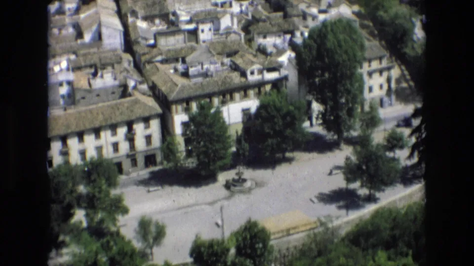 Elevated 1972 Spain view over clustered rooftops, pale residential buildings, trees, and a small plaza fountain in bright daylight.