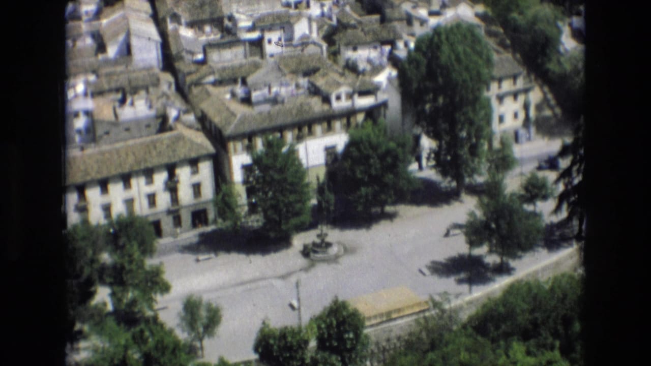 1972 Spain 8mm frame showing an elevated view over a sunlit town square with pale buildings, clay-tiled roofs, trees, and a small central fountain.