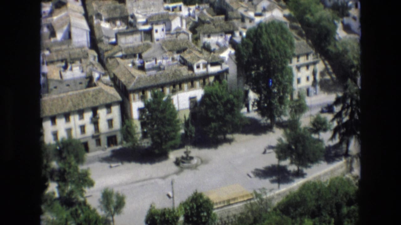 Elevated town-square view with buildings, trees, roofs, and a central fountain seen through dark film borders.