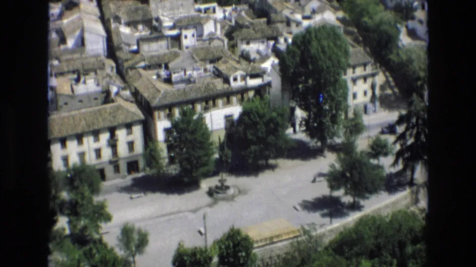 1972 Spain home-movie frame looking down on a town square bordered by traditional buildings, trees, clay-tiled roofs, and a small fountain.